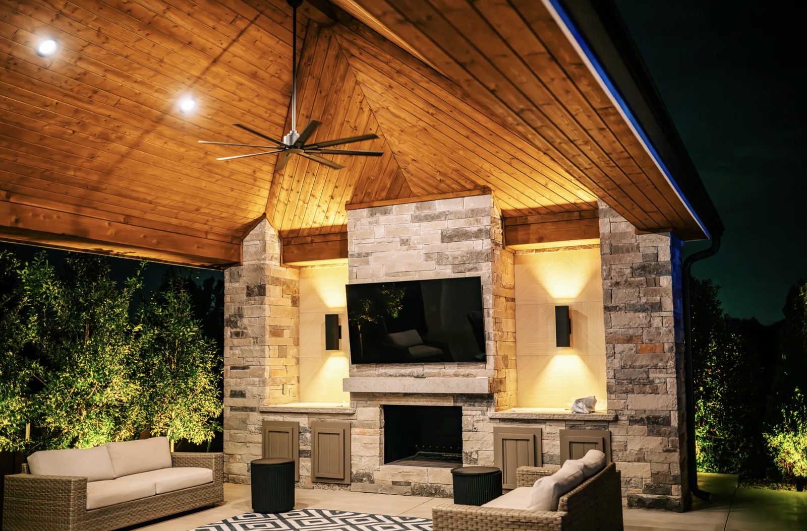 Covered outdoor living pavilion at night with vaulted cedar ceiling, stacked stone fireplace, television, and warm sconce lighting
