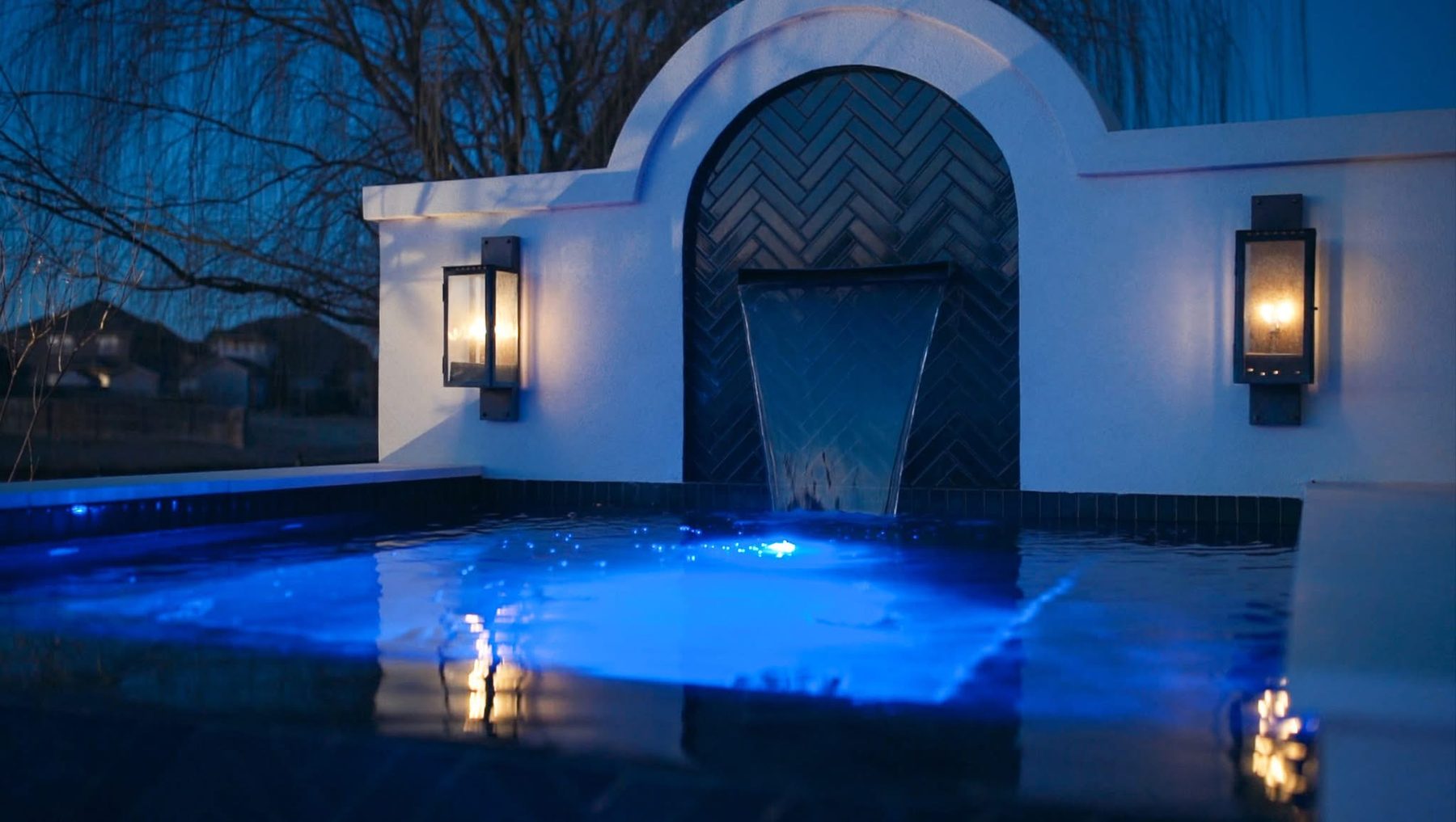 Custom gunite pool at dusk with an arched stucco water feature wall, herringbone tile, and lantern sconces