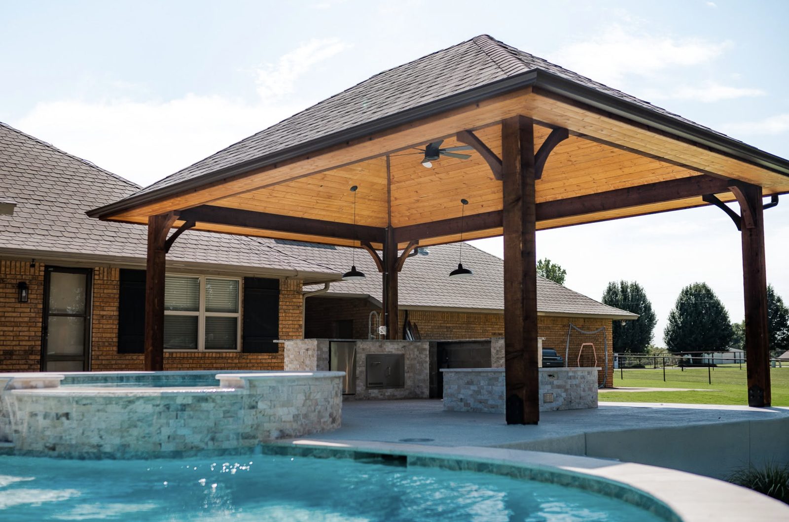 Timber pavilion with vaulted wood ceiling covering a stone outdoor kitchen beside a pool and raised spa