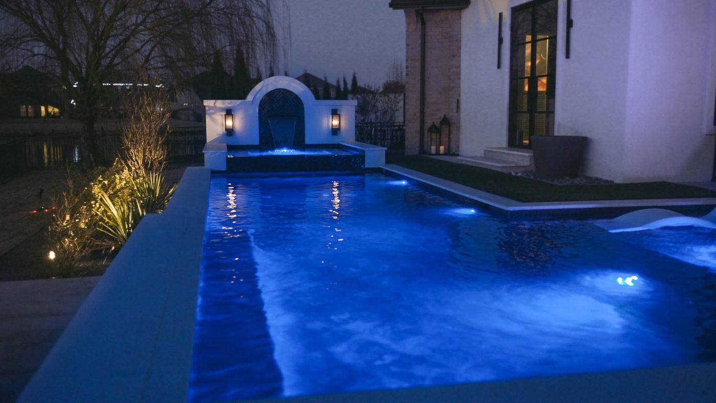 Custom gunite pool glowing blue at night beside the home, with landscape lighting on the surrounding planting