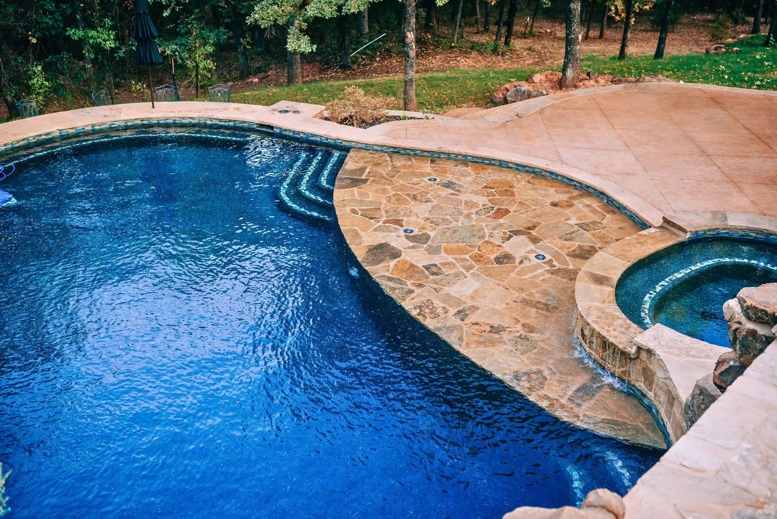 Freeform gunite pool with a flagstone sun shelf, glass tile steps, and raised spa on a wooded lot