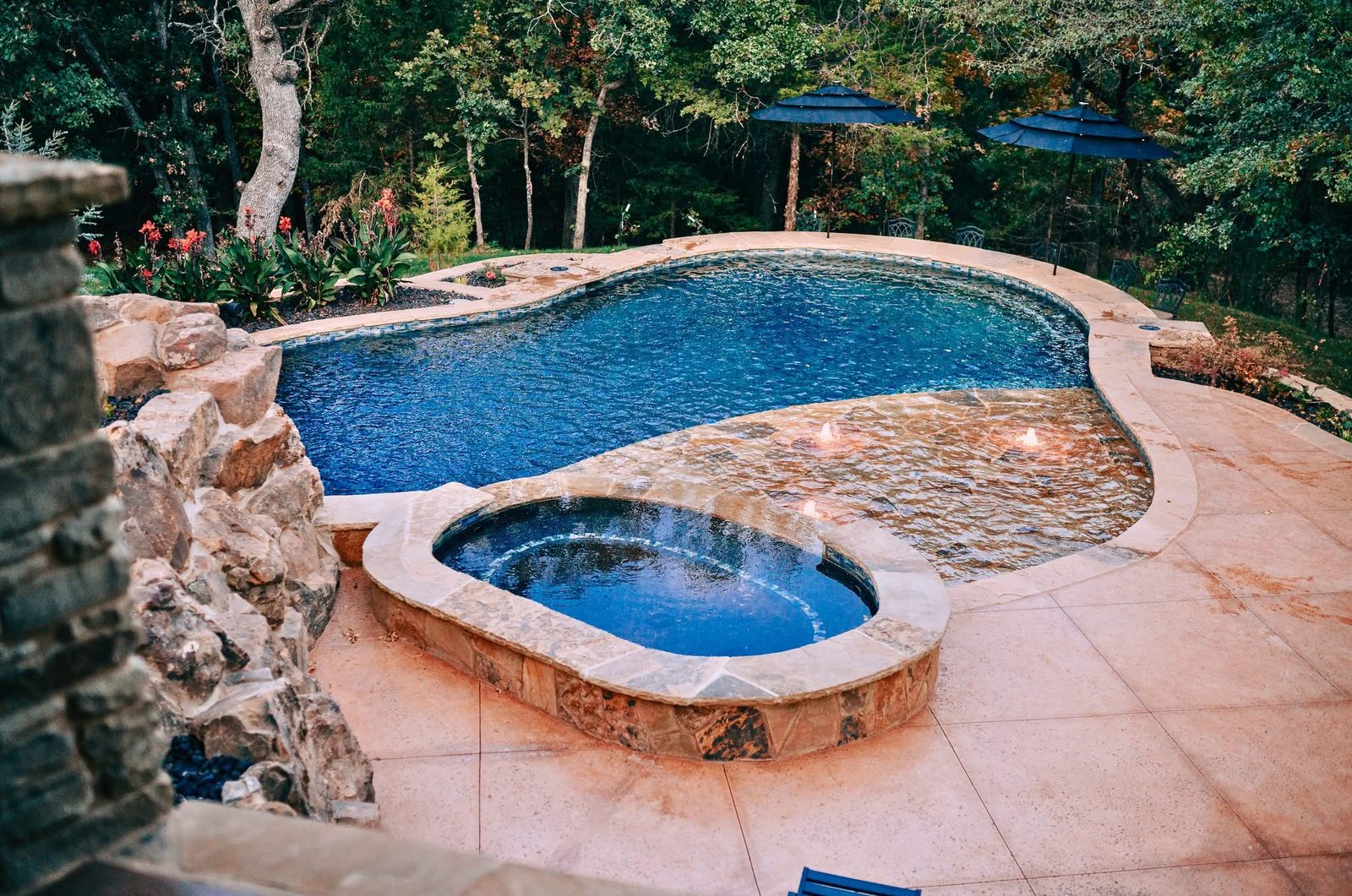 Freeform pool with raised oval spa and fire bowls burning on a submerged stone ledge, surrounded by stamped concrete decking