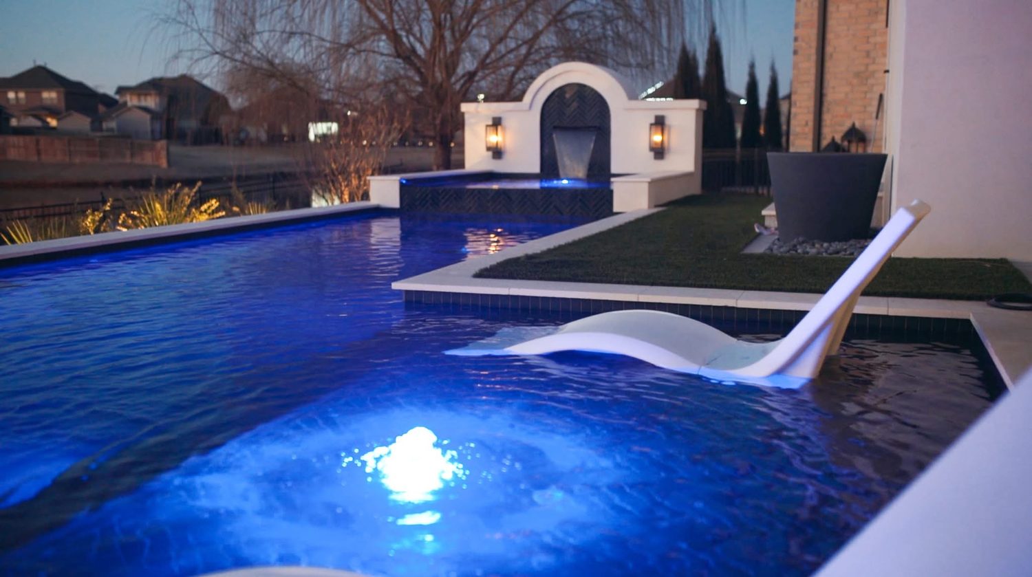 Evening view of a custom pool with an in-water tanning ledge lounger, raised spa with fountain wall, and turf surround