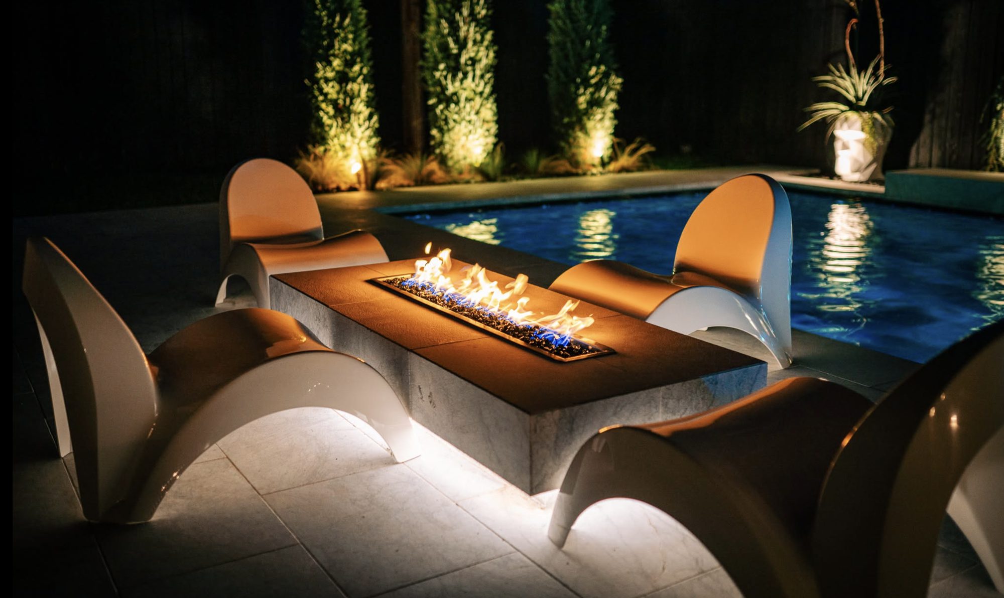 Linear gas fire feature and sculpted lounge chairs beside a lit pool at night, with uplit cypress landscaping