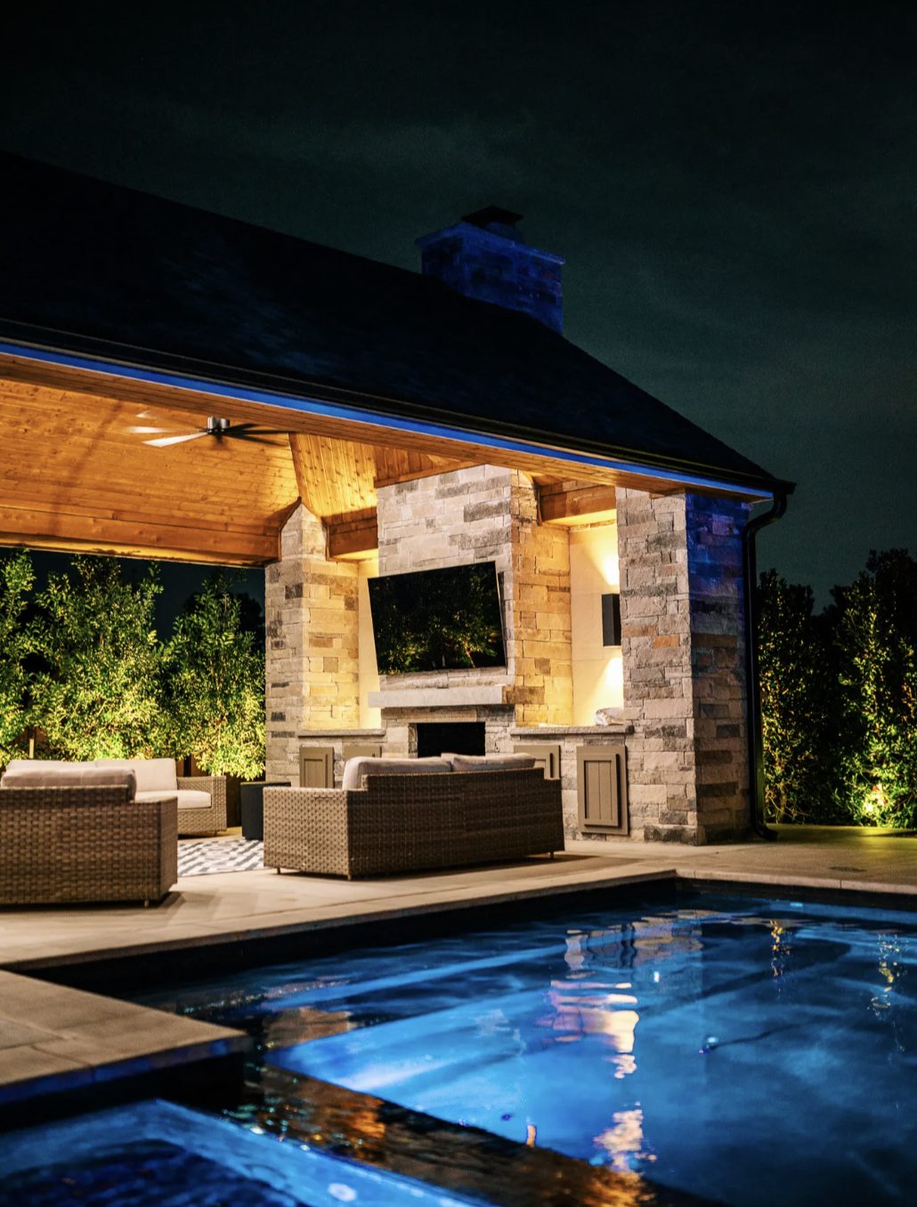 Poolside pavilion at night with a stone fireplace, mounted television, and lounge seating under a wood ceiling