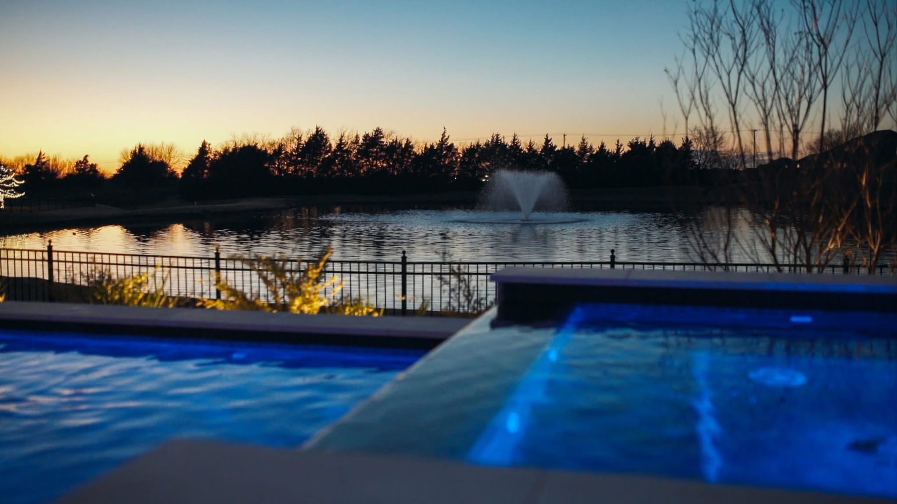 Pool and raised spa at sunset overlooking a pond with a fountain beyond an iron fence