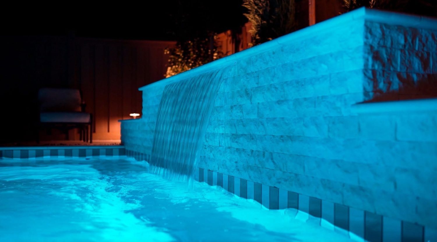 Sheer descent waterfall spilling over a stacked stone wall into a pool lit teal at night