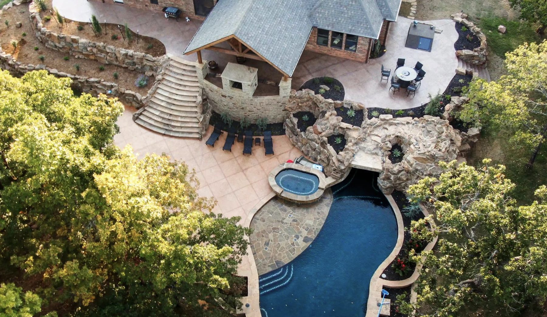 Aerial view of a resort-style estate backyard with a natural stone grotto pool, spa, curved stone staircase, and covered pavilion